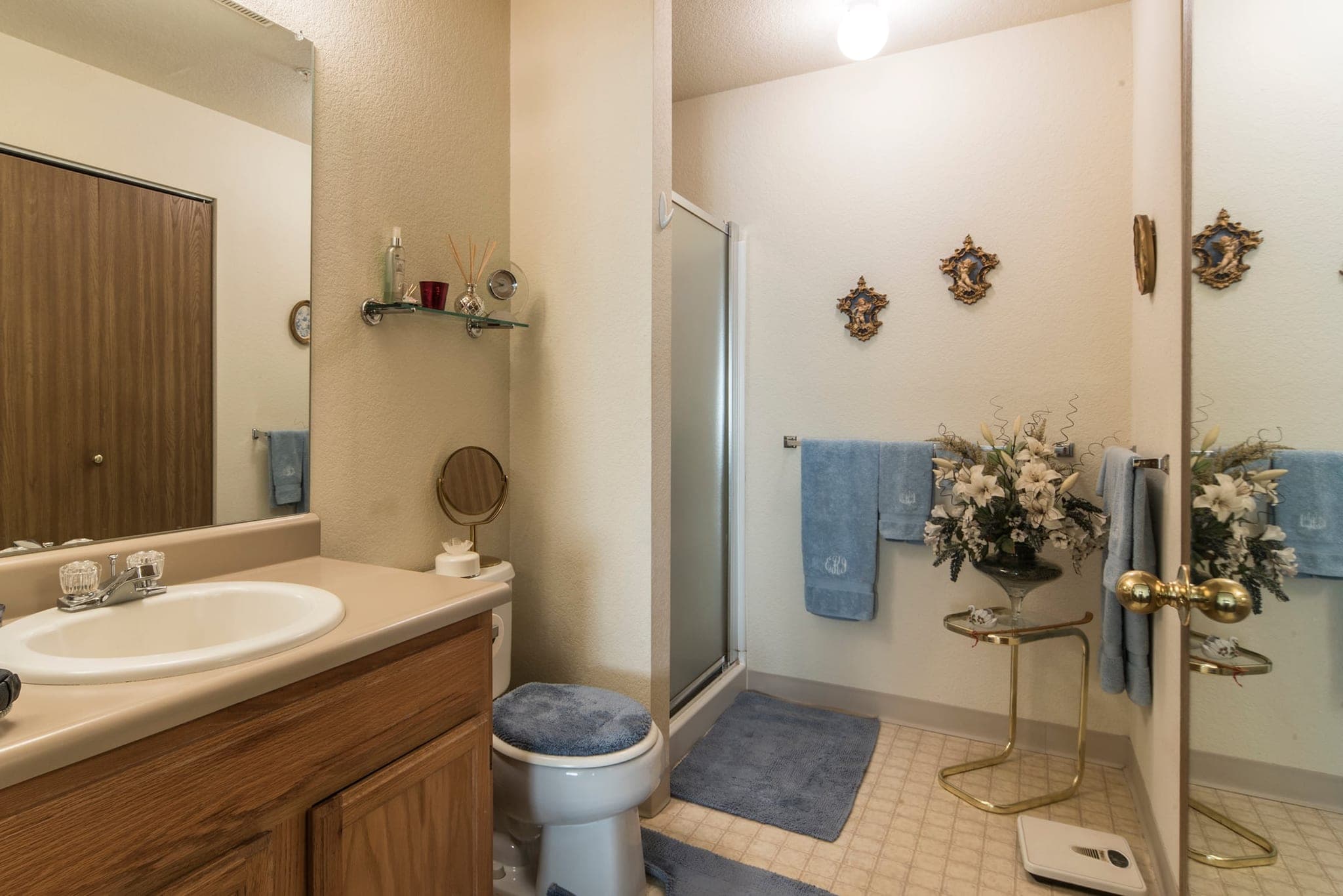 A bathroom in use, with an oak vanity and a wide mirror, a step-in shower stall with a glass door, folded towels on a rail and a vase of flowers on the shelf.