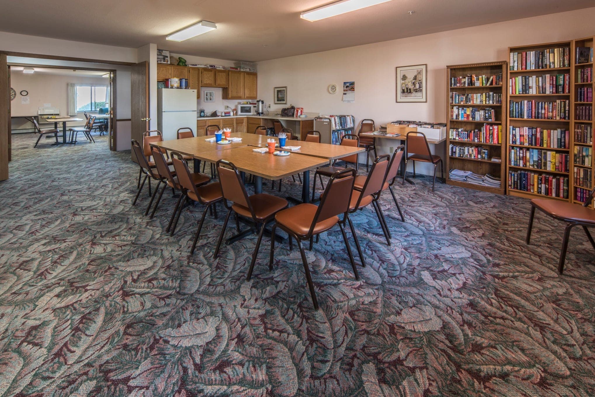 The community library: two tall bookcases filled with books, long tables set with chairs for a dozen people, a kitchenette along the far wall and a bright seating area beyond.